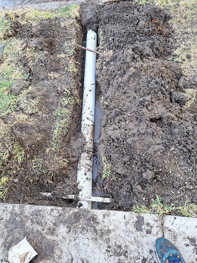 Underground Drainage Pipe Installation - Emerson Pro Services White PVC drainage pipe installed in a narrow trench beside a concrete walkway.