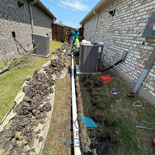 Technician installing an underground drainage pipe in a trench between two brick homes.